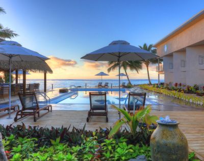 Outdoor pool area overlooking the ocean at sunset, with deck chairs, umbrellas, and lush greenery, conveying a serene and tropical resort ambiance.