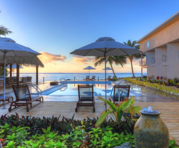 Outdoor pool area overlooking the ocean at sunset, with deck chairs, umbrellas, and lush greenery, conveying a serene and tropical resort ambiance.