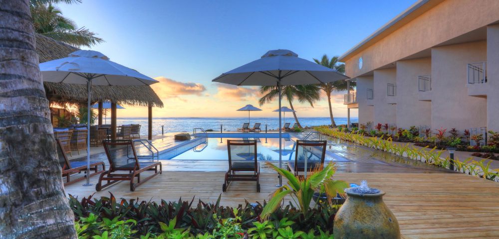 Outdoor pool area overlooking the ocean at sunset, with deck chairs, umbrellas, and lush greenery, conveying a serene and tropical resort ambiance.