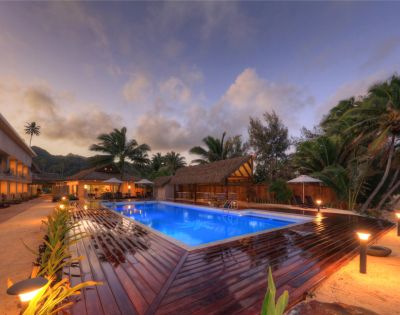Tropical resort at dusk with a glowing blue pool, surrounded by palm trees and thatched huts. Warm lights illuminate pathways and a serene beach lies nearby.