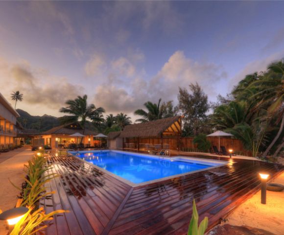 Tropical resort at dusk with a glowing blue pool, surrounded by palm trees and thatched huts. Warm lights illuminate pathways and a serene beach lies nearby.