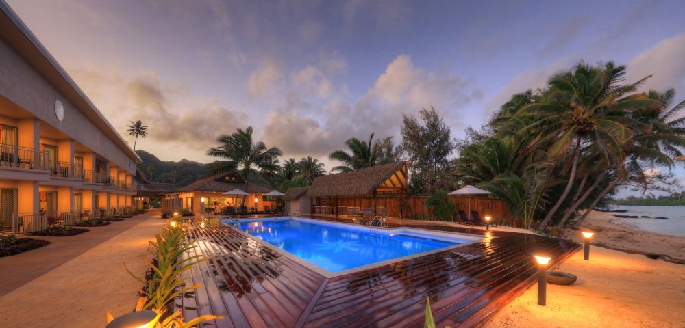 Tropical resort at dusk with a glowing blue pool, surrounded by palm trees and thatched huts. Warm lights illuminate pathways and a serene beach lies nearby.