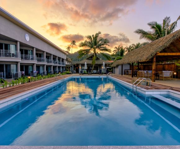 Sunset view of a serene hotel pool with a palm tree reflection. Modern building on the left, thatched-roof seating on the right, tropical ambiance.