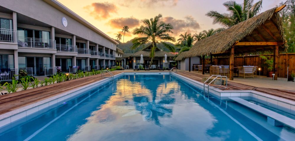 Sunset view of a serene hotel pool with a palm tree reflection. Modern building on the left, thatched-roof seating on the right, tropical ambiance.