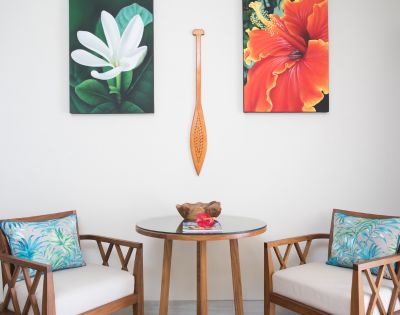 Cozy seating area with a round glass table, two wooden chairs with floral cushions. Above, a white flower and a red-orange flower painting flank a wooden paddle.