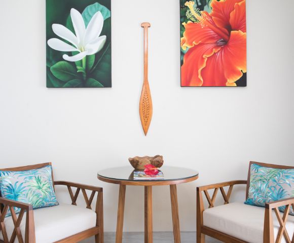Cozy seating area with a round glass table, two wooden chairs with floral cushions. Above, a white flower and a red-orange flower painting flank a wooden paddle.