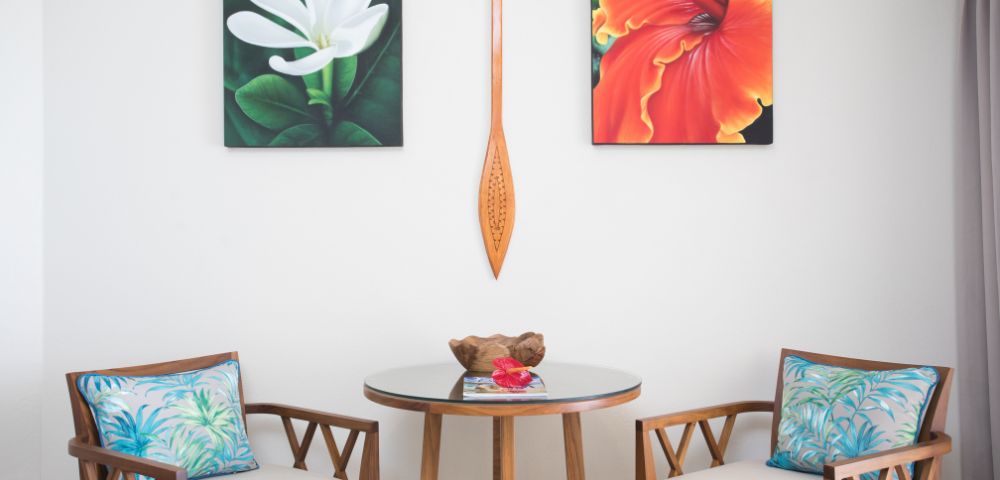Cozy seating area with a round glass table, two wooden chairs with floral cushions. Above, a white flower and a red-orange flower painting flank a wooden paddle.