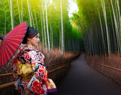 A woman in a floral kimono holds a red parasol, standing on a path lined with tall bamboo and dried grass, creating a serene and peaceful atmosphere.