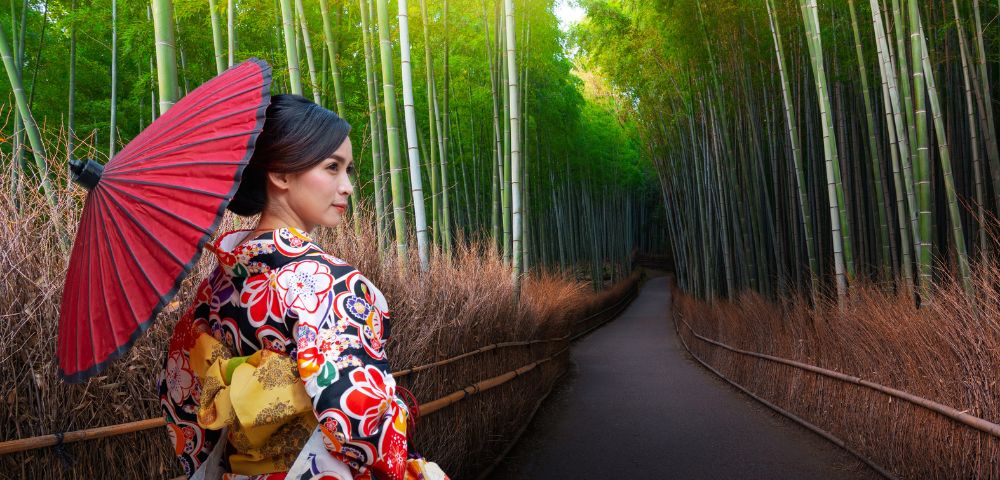 A woman in a floral kimono holds a red parasol, standing on a path lined with tall bamboo and dried grass, creating a serene and peaceful atmosphere.