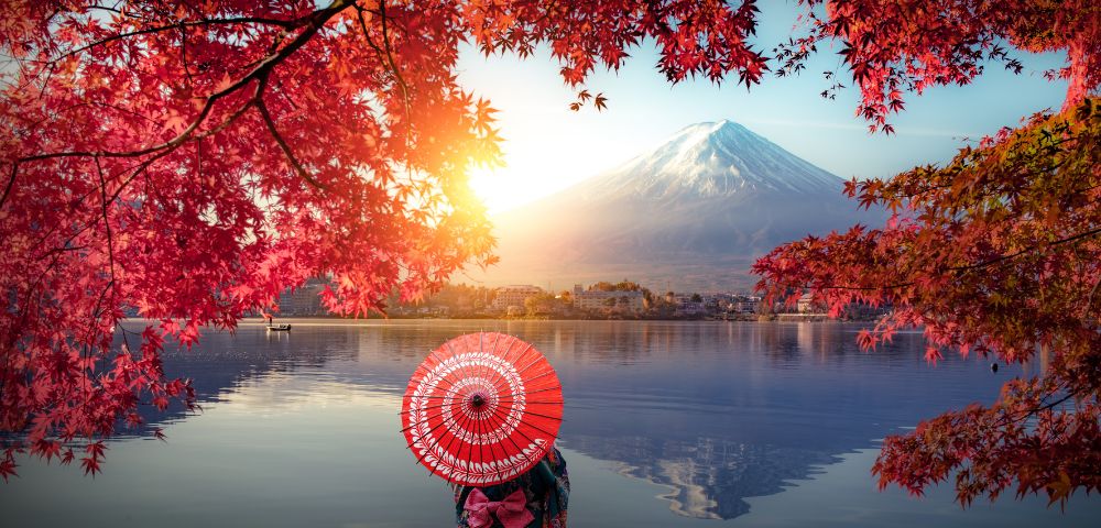 A person with a red parasol stands by a tranquil lake, facing Mount Fuji. Autumn leaves frame the scene, and the sun casts a warm glow, creating a serene atmosphere.