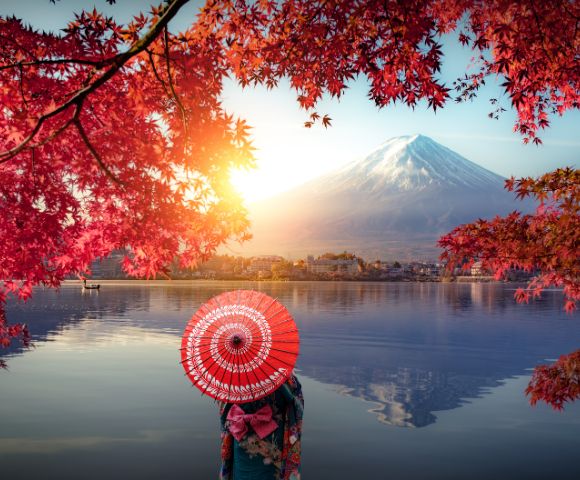 A person with a red parasol stands by a tranquil lake, facing Mount Fuji. Autumn leaves frame the scene, and the sun casts a warm glow, creating a serene atmosphere.