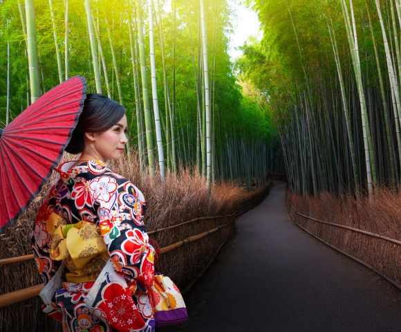 A woman in a floral kimono holds a red parasol, standing on a path lined with tall bamboo and dried grass, creating a serene and peaceful atmosphere.