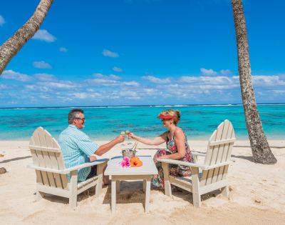 A couple sits on wooden chairs, clinking glasses on a sandy beach. The turquoise ocean and a clear blue sky create a tranquil, tropical atmosphere.