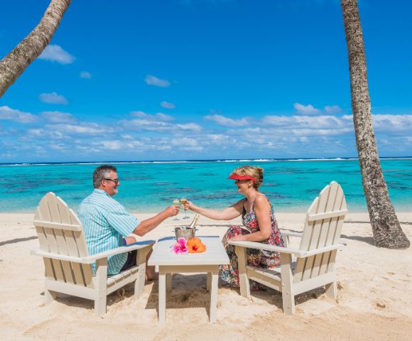 A couple sits on wooden chairs, clinking glasses on a sandy beach. The turquoise ocean and a clear blue sky create a tranquil, tropical atmosphere.