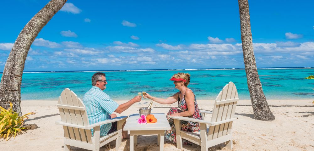 A couple sits on wooden chairs, clinking glasses on a sandy beach. The turquoise ocean and a clear blue sky create a tranquil, tropical atmosphere.