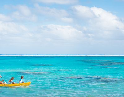 A serene beach scene with two people kayaking in a bright yellow kayak on crystal-clear turquoise waters under a partly cloudy sky, conveying tranquility.