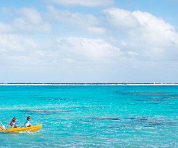 A serene beach scene with two people kayaking in a bright yellow kayak on crystal-clear turquoise waters under a partly cloudy sky, conveying tranquility.