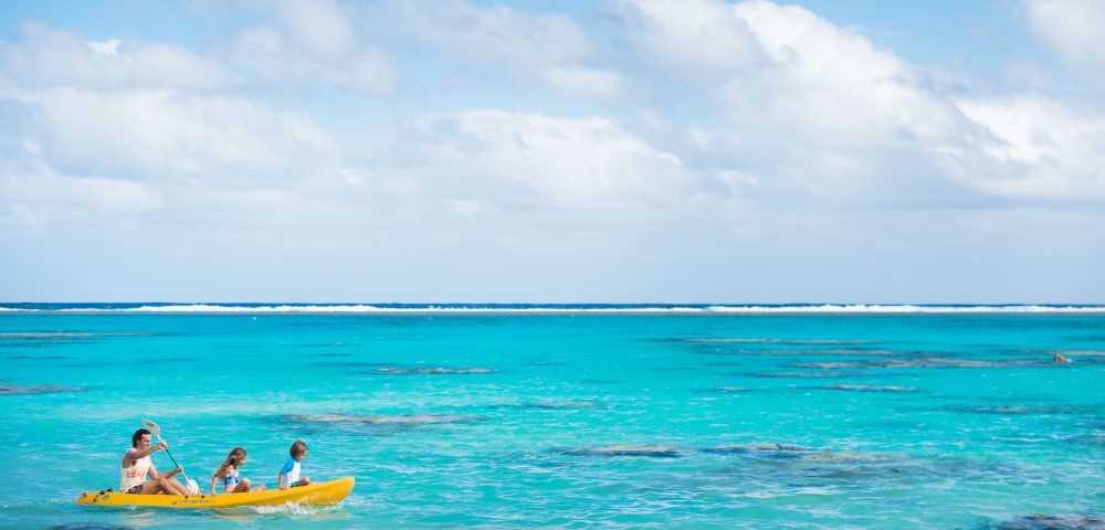 A serene beach scene with two people kayaking in a bright yellow kayak on crystal-clear turquoise waters under a partly cloudy sky, conveying tranquility.