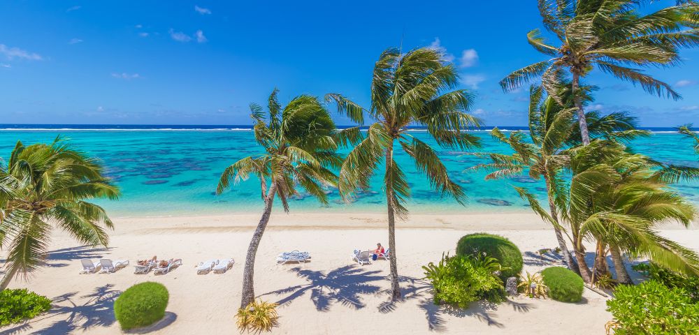 Tropical beach scene with turquoise water, white sand, and swaying palm trees. Clear blue sky and vibrant lush greenery create a serene, relaxing atmosphere.