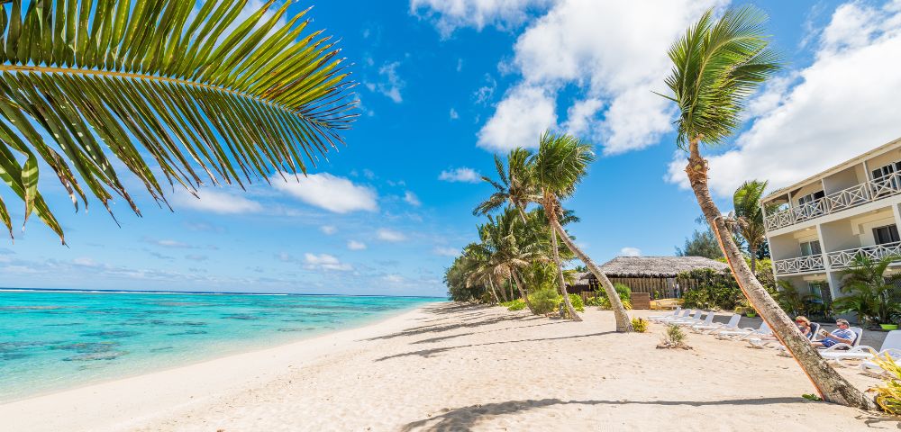 Sunny tropical beach with white sand, turquoise sea, and swaying palm trees. A building with balconies is visible, adding a serene, inviting vibe.
