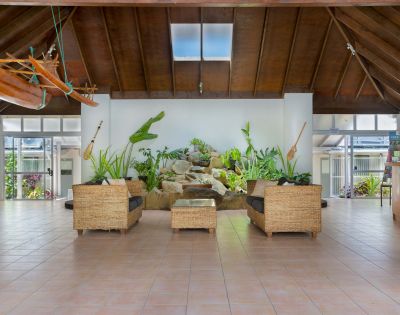 A spacious tropical atrium with wicker seating, vibrant plants, and wall-mounted paddles. A skylight and wooden ceiling create an inviting, airy ambiance.