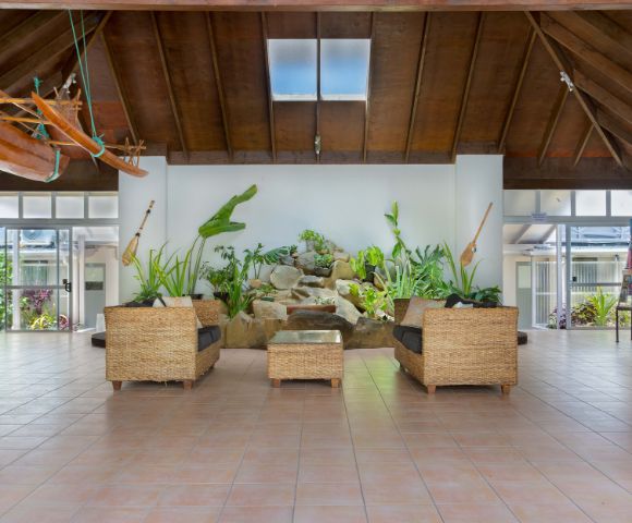 A spacious tropical atrium with wicker seating, vibrant plants, and wall-mounted paddles. A skylight and wooden ceiling create an inviting, airy ambiance.