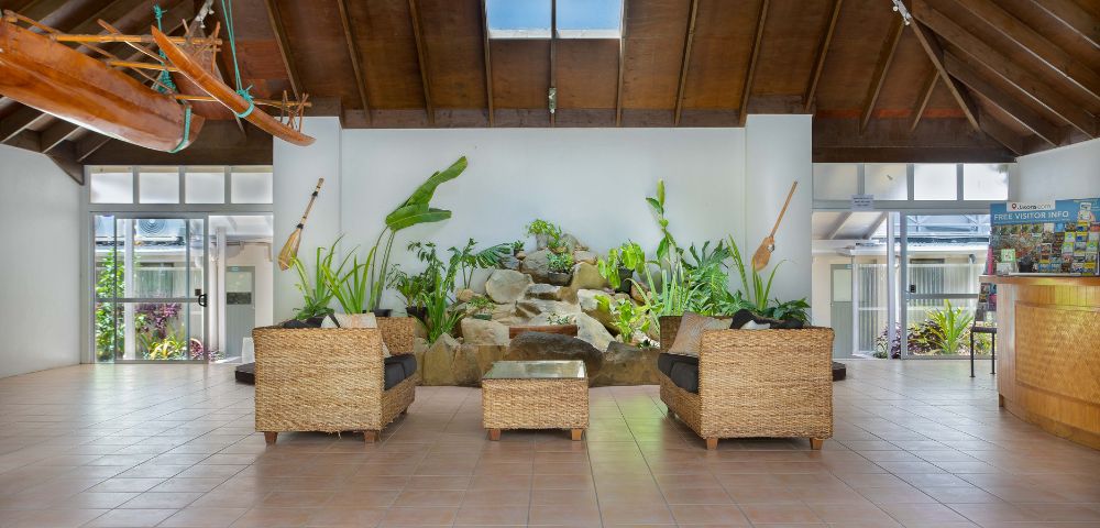 A spacious tropical atrium with wicker seating, vibrant plants, and wall-mounted paddles. A skylight and wooden ceiling create an inviting, airy ambiance.