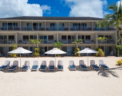 A beachfront hotel with three stories overlooks a sandy shore lined with empty loungers and white umbrellas under a bright blue sky and scattered clouds.