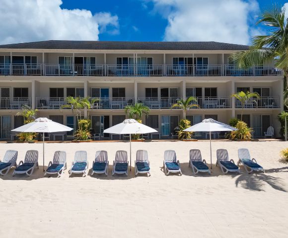 A beachfront hotel with three stories overlooks a sandy shore lined with empty loungers and white umbrellas under a bright blue sky and scattered clouds.