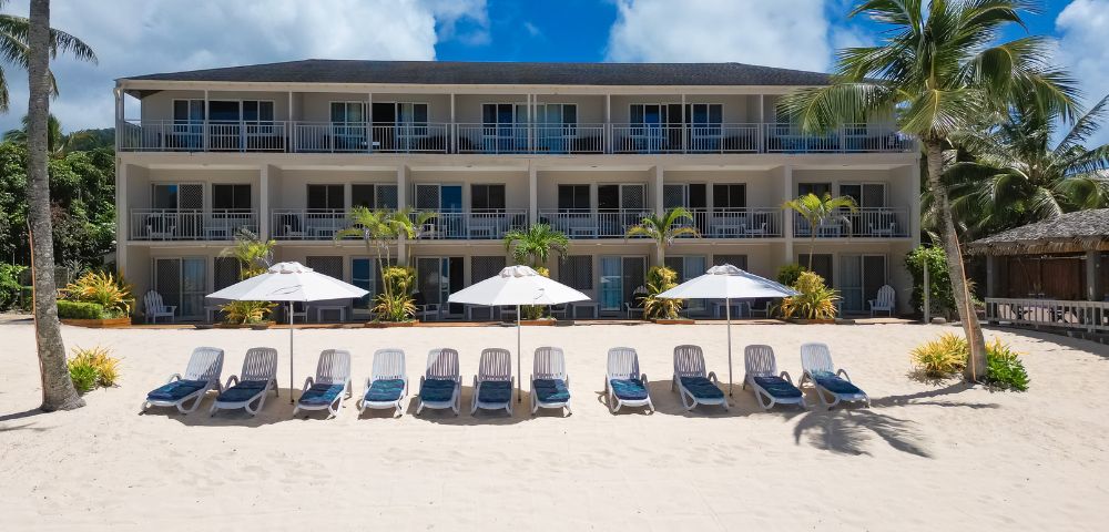 A beachfront hotel with three stories overlooks a sandy shore lined with empty loungers and white umbrellas under a bright blue sky and scattered clouds.