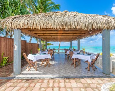 Tropical beachside gazebo with a thatched roof and open sides, featuring tables set for dining. Ocean and palm trees visible in the background.