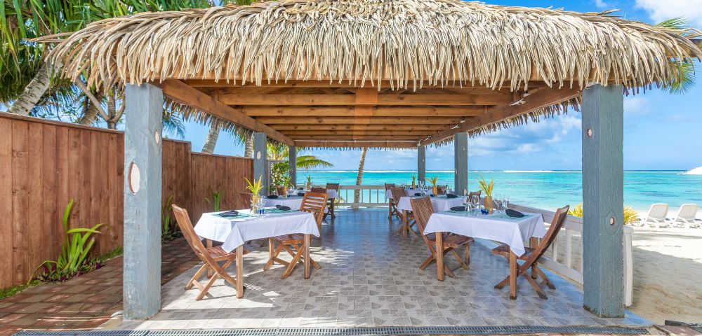 Tropical beachside gazebo with a thatched roof and open sides, featuring tables set for dining. Ocean and palm trees visible in the background.