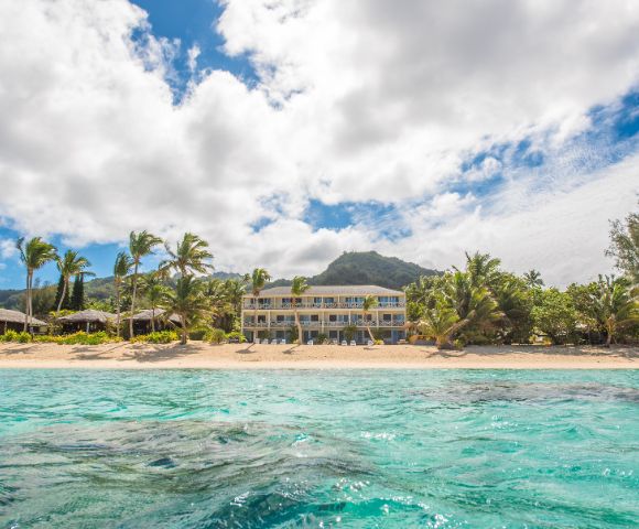 Tropical beach scene with turquoise water, sandy shore, palm trees, and a large resort building. Background features lush mountains under a partly cloudy sky.