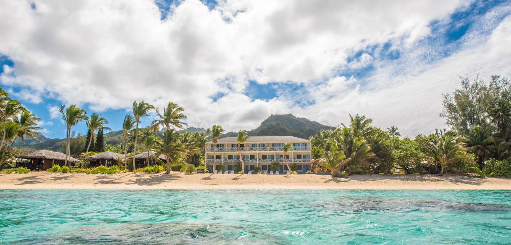 Tropical beach scene with turquoise water, sandy shore, palm trees, and a large resort building. Background features lush mountains under a partly cloudy sky.