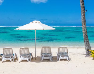Five white lounge chairs under a large umbrella face a tranquil turquoise sea on a sandy beach, evoking a serene and relaxing atmosphere.
