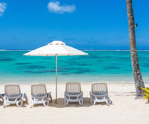 Five white lounge chairs under a large umbrella face a tranquil turquoise sea on a sandy beach, evoking a serene and relaxing atmosphere.