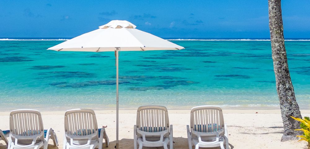 Five white lounge chairs under a large umbrella face a tranquil turquoise sea on a sandy beach, evoking a serene and relaxing atmosphere.