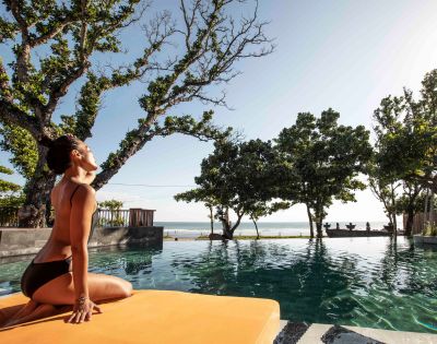 A woman in a black bikini sits on an orange mat by a pool, stretching with serene expression. Lush trees surround, and the ocean is visible in the background.