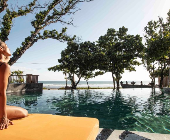 A woman in a black bikini sits on an orange mat by a pool, stretching with serene expression. Lush trees surround, and the ocean is visible in the background.