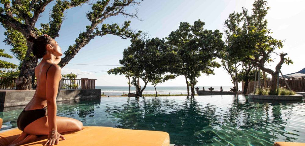 A woman in a black bikini sits on an orange mat by a pool, stretching with serene expression. Lush trees surround, and the ocean is visible in the background.