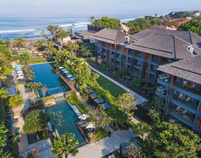 Aerial view of a tropical resort by the ocean, featuring a wide pool surrounded by lush greenery and lounge chairs, with waves in the background.