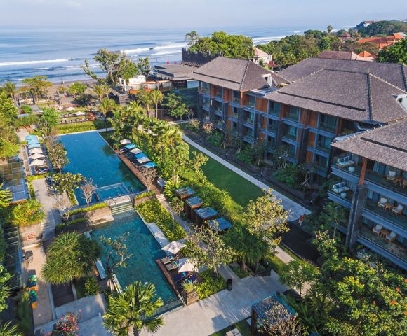 Aerial view of a tropical resort by the ocean, featuring a wide pool surrounded by lush greenery and lounge chairs, with waves in the background.