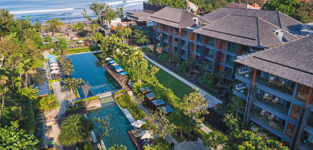 Aerial view of a tropical resort by the ocean, featuring a wide pool surrounded by lush greenery and lounge chairs, with waves in the background.
