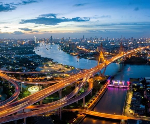 Aerial view of a vibrant cityscape at dusk, featuring illuminated highways and bridges over a river, surrounded by urban buildings and glowing lights.