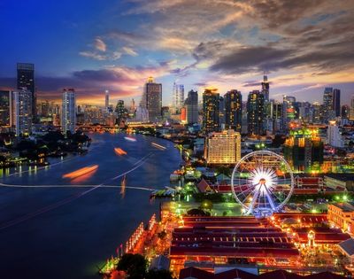 A vibrant cityscape at dusk with a river, modern skyscrapers, a Ferris wheel, and colorful lights reflecting on the water, conveying an energetic urban vibe.