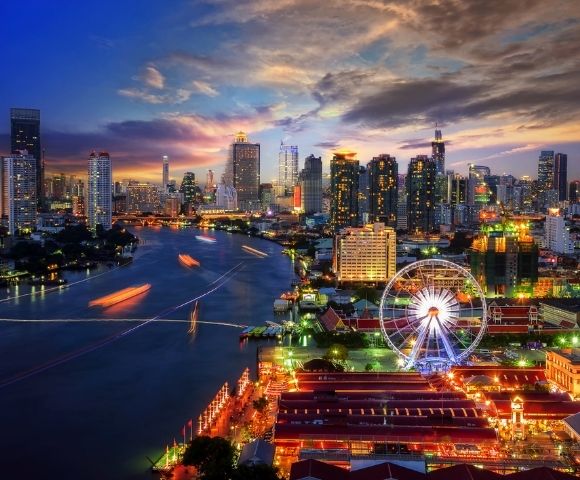 A vibrant cityscape at dusk with a river, modern skyscrapers, a Ferris wheel, and colorful lights reflecting on the water, conveying an energetic urban vibe.