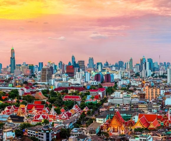 Skyline of a bustling city at sunset, with modern skyscrapers and traditional red-roofed temples. The vibrant sky adds warmth and a sense of tranquility.