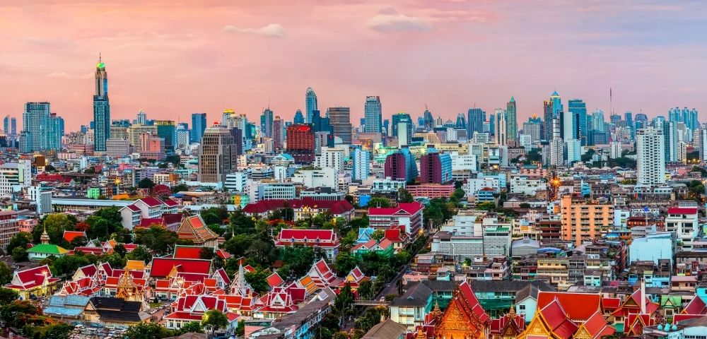 Skyline of a bustling city at sunset, with modern skyscrapers and traditional red-roofed temples. The vibrant sky adds warmth and a sense of tranquility.