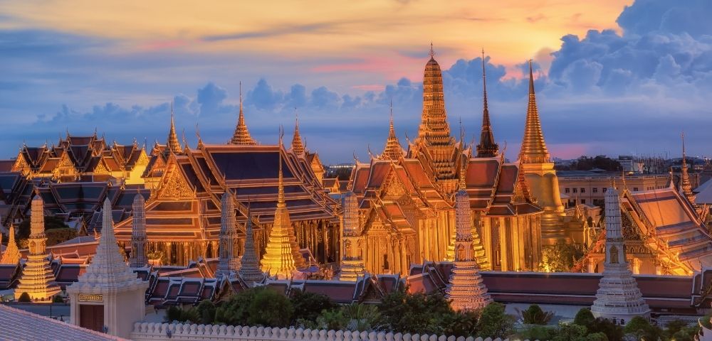 The image shows Bangkok's Grand Palace illuminated at sunset, with intricate spires and roofs against a vibrant sky, creating a serene, majestic atmosphere.