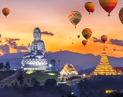 Sunset over Chiang Rai with a giant white Buddha statue and golden temple, surrounded by colorful hot air balloons in a serene, majestic landscape.